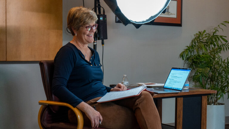 Photo of Brenda VanLengen with a notebook on her lap and a computer by her side interviewing a subject for the docuseries.